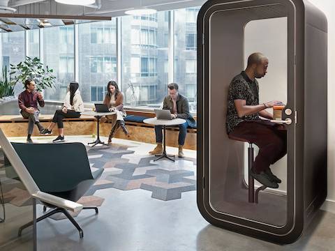 Una persona sentada en un taburete dentro de una cabina telefónica Framery O para oficinas. Cerca, cuatro personas sentadas conversan y trabajan en portátiles en un asiento de ventana.