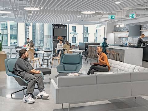 A person sits on an blue Striad Lounge Chair talking to a person sitting on a grey Tuxedo Sofa in a lounge space. In the background, people gather in a cafe setting. 