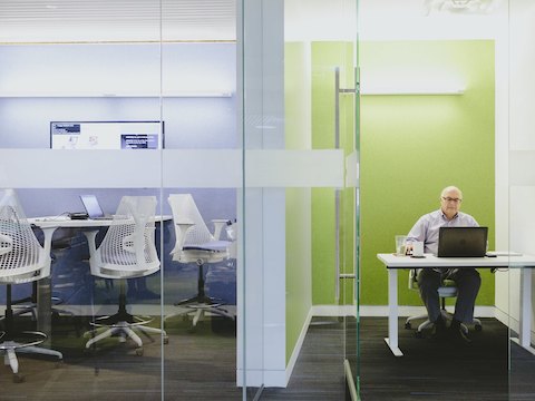 A man sits at his desk inside of an enclosed office space with glass walls. 