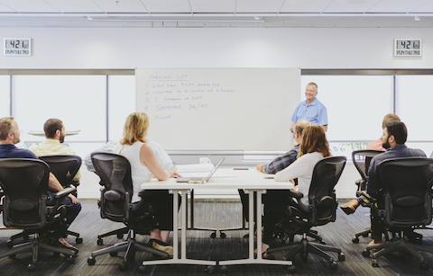 Medewerkers zitten in Aeron-stoelen en luisteren naar een presentatie in een vergaderruimte.