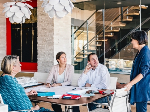 Office staff talk while seated at a table within an open meeting area. 