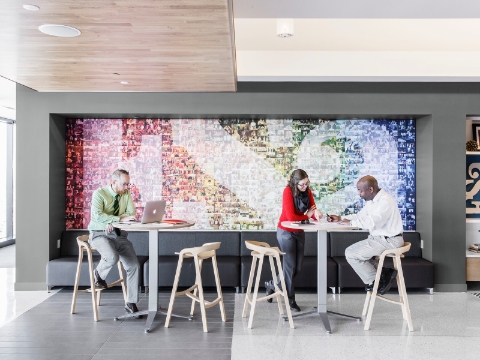 Employees talk and work at tall desks while seated on stools in an open gathering area. 