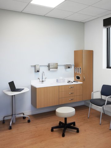 Patient exam room with Mora casework in Oak on Ash and a white solid surface top, an Intent mobile table with a white top and silver base, two Verus side chairs in a dark blue upholstery with a Nemschoff Riva bench in a dark blue upholstery, and a physician's stool in a soft white seat and black base.