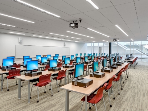 Computers sit atop several rows of desk and chairs within a university building. Computers sit atop several rows of desk and chairs within a university building.
