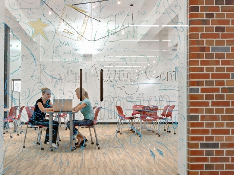 Two women look at a computer screen while seated inside of a glass-walled room. Two women look at a computer screen while seated inside of a glass-walled room.