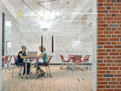 Two women look at a computer screen while seated inside of a glass-walled room. 