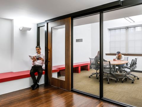 A man reads a textbook while waiting outside of a glass-walled meeting room. A man reads a textbook while waiting outside of a glass-walled meeting room.