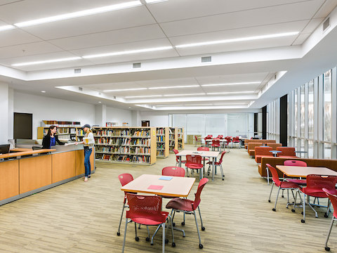 Two women talk inside of a library furnished with red Caper chairs. 