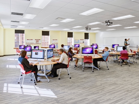 Students sit at round computer tables inside of a university classroom. 