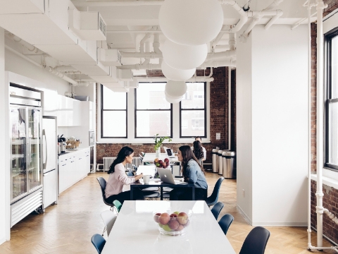 Two women sit and talk inside of an office cafe area full of natural light.