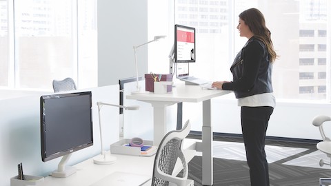 A woman types on a laptop at a standing desk near a window. A woman types on a laptop at a standing desk near a window.