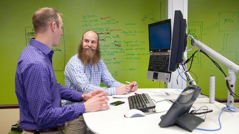 Two men converse in front of an extended computer monitor setup. 