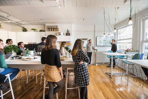 Office staff listen to a co-worker's presentation in a multi-purpose room with windows. 