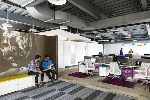 Two office employees talk at a bench seating area away from their workstations. 