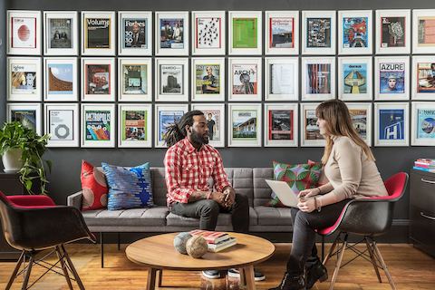 Two people talk in a lounge area with framed magazine covers and ads on the wall.