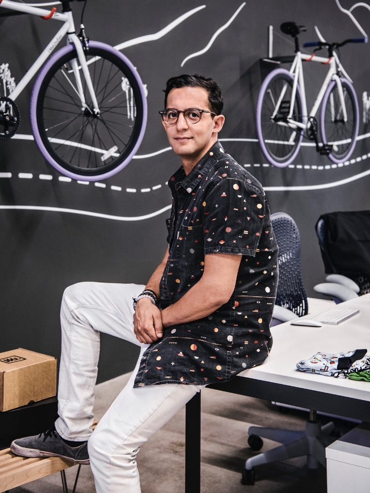 A man sits on a desk, with bicycles as decor on the wall behind him. 