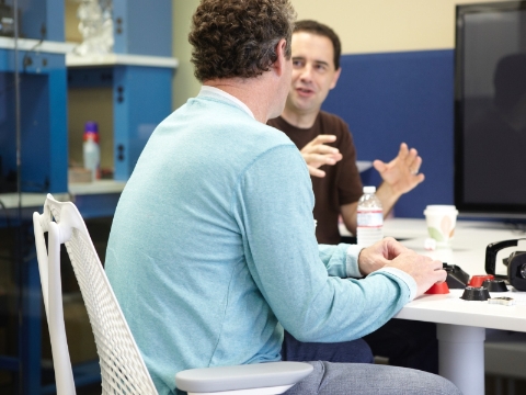 Two men converse as they sit at a table inside an office. Two men converse as they sit at a table inside an office.