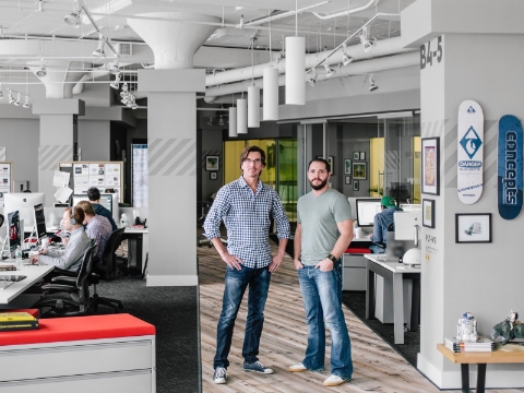 Two men pose in an office space while others work at their desks in the background.