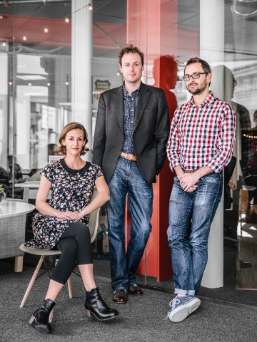 Three people pose in front of an office space with glass walls. 