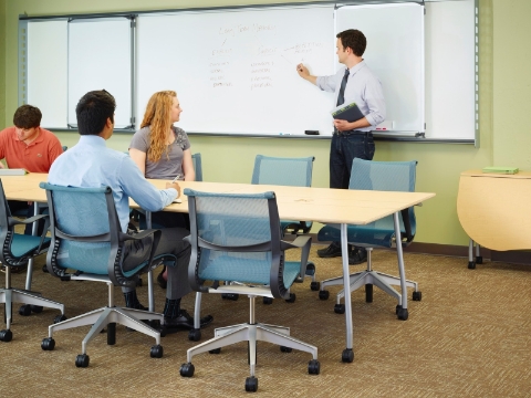 Students observe an instructor while seated at Mobile tables inside a classroom. 