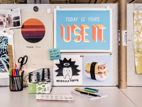 A bulletin board full of notes and pictures sits behind a desk space. 