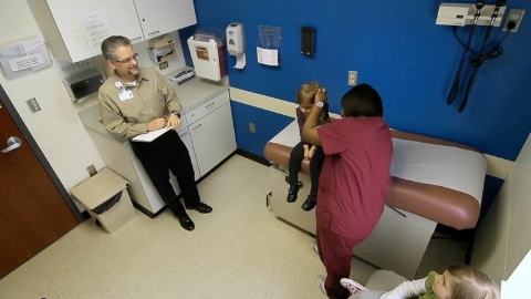 A nurse examines a child's ear while a man with a clipboard looks on. Select to go to a case study featuring Spectrum Health.