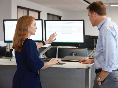 Twee mensen praten terwijl ze naar een monitor op een sta-bureau kijken.