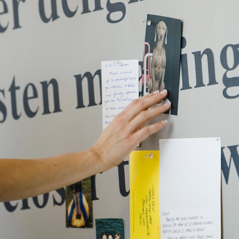 A close up of a hand tacking a photo of a dog to a board covered in a fabric with black words on it and with other notes and photos posted on it.