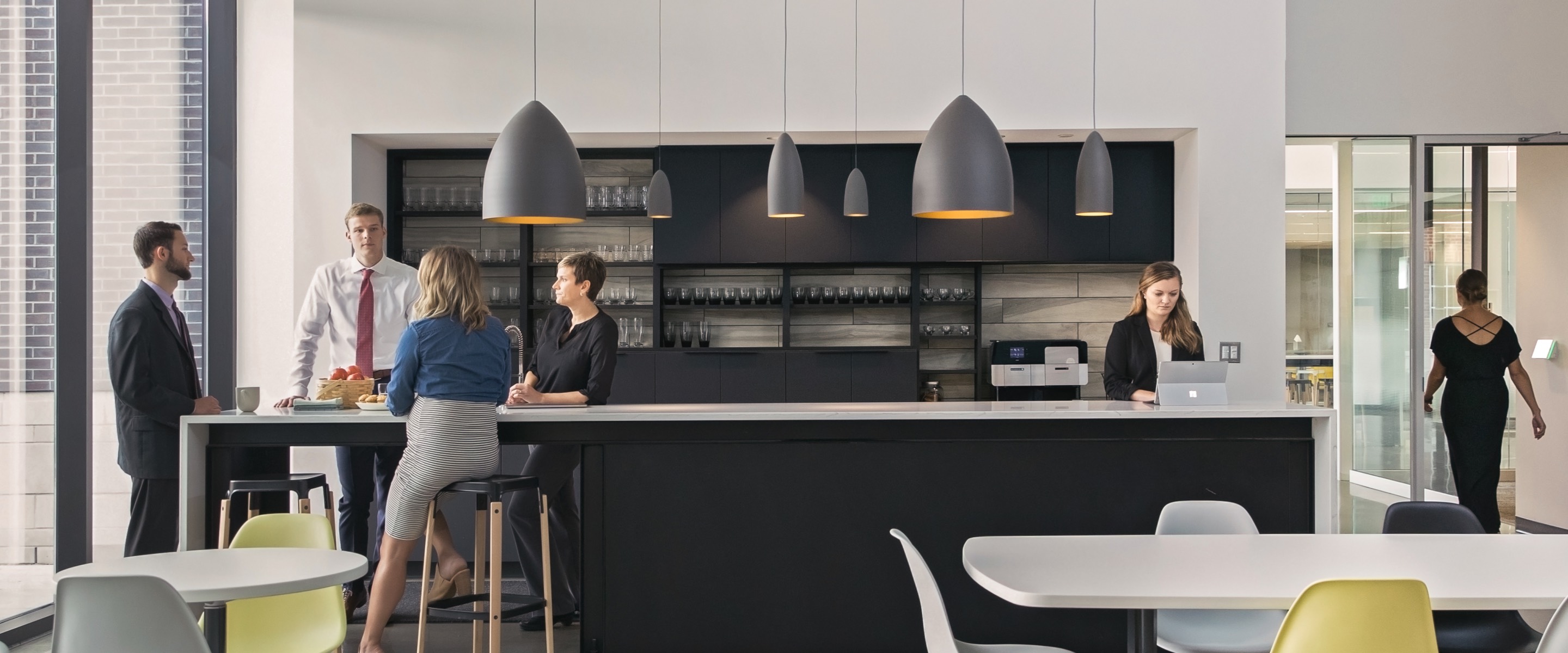 People socializing at a long bar in a break room with white tables and yellow and white Eames Shell Chairs.