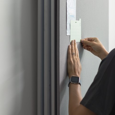 A close-up of a person's hand posting a note on a gray tackboard.