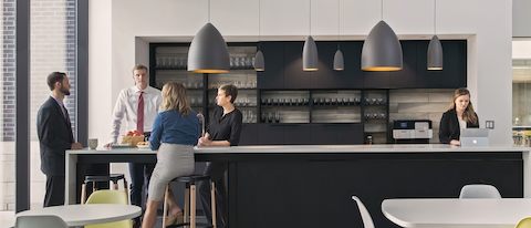 People socializing at a long bar in a break room with white tables and yellow and white Eames Shell Chairs.