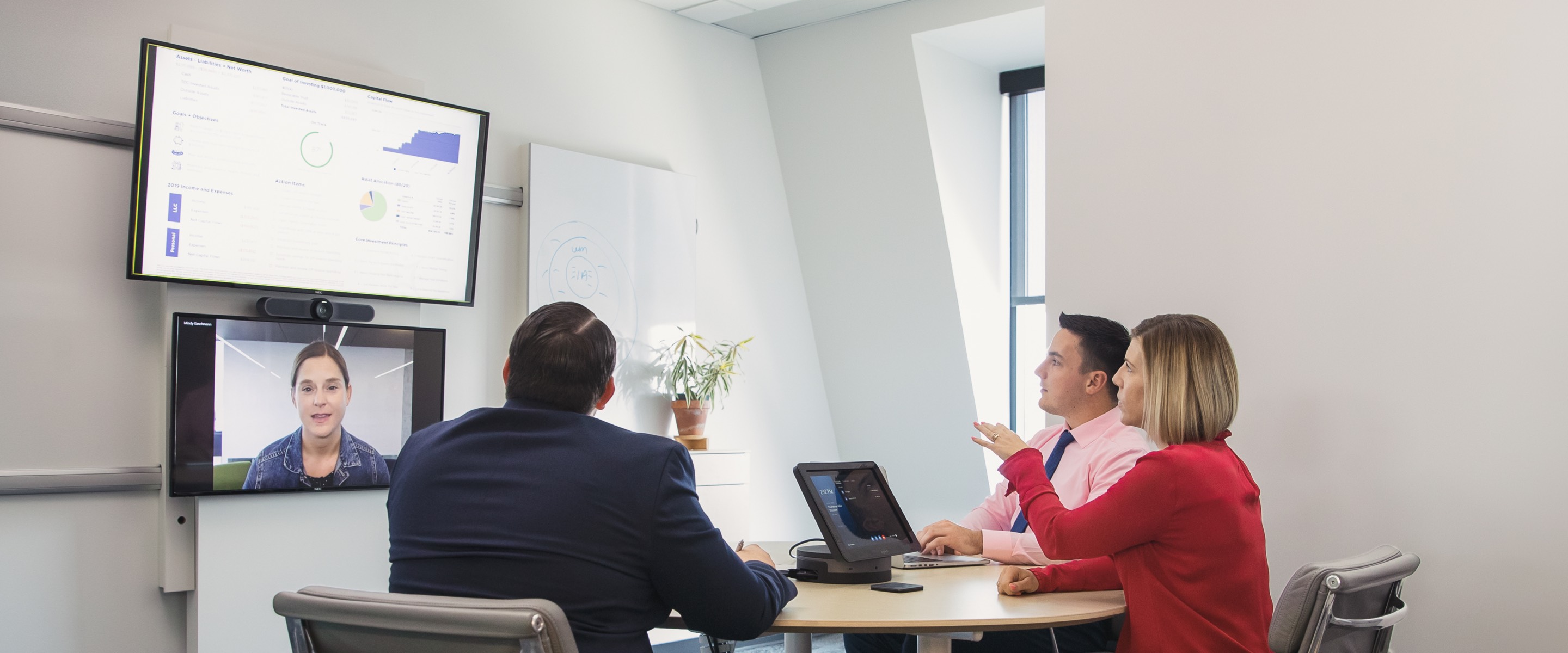 Three people sitting in gray Eames Soft Pad Chairs talking with a remote colleague whose face appears on a large monitor on the wall.