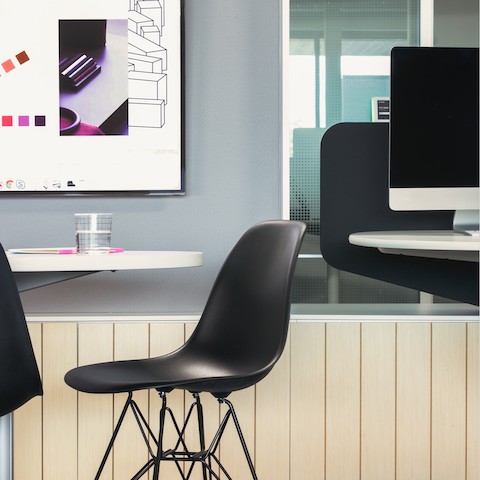 A side view of a black Eames Shell chair next to a white table with a monitor in the background. A side view of a black Eames Shell chair next to a white table with a monitor in the background.