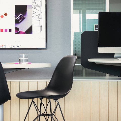 A side view of a black Eames Shell chair next to a white table with a monitor in the background.