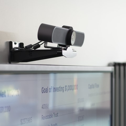 A black video camera affixed to a wall above a screen that's used for video conferencing.