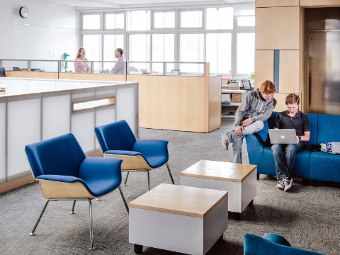 Two students look at a laptop screen together as they sit on a blue couch. Two students look at a laptop screen together as they sit on a blue couch.