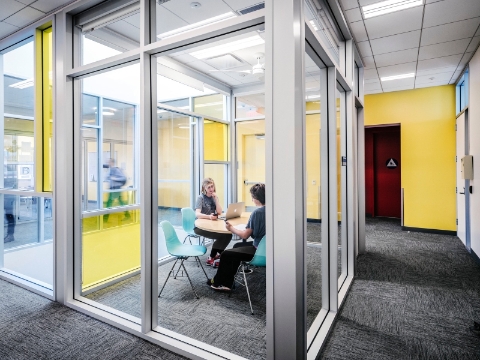 Two people sit at a desk inside of an enclosed space with glass walls. Two people sit at a desk inside of an enclosed space with glass walls.