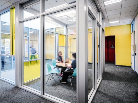 Two people sit at a desk inside of an enclosed space with glass walls. 