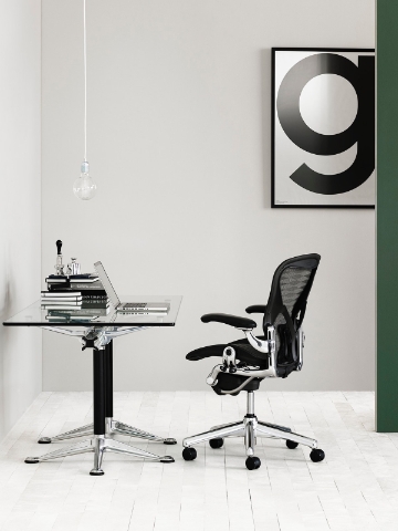 An Aeron chair sits behind a glass top desk with a minimalist set up. 