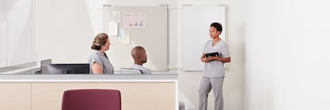 Three clinicians talking in a care team work area created by a Commend Nurses Station in light ash with glass to the ceiling.