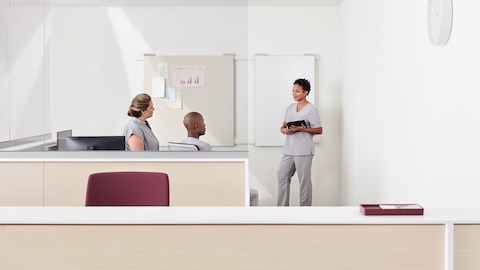 Three clinicians talking in a care team work area created by a Commend Nurses Station in light ash with glass to the ceiling.