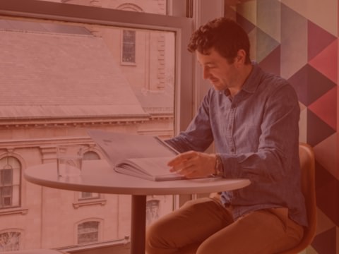 A person sits at a high top desk and reads a book. The surrounding office is bright and airy, and is outfitted with design elements that reflect the company's brand.