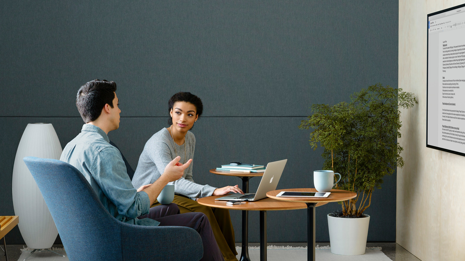 Two people meeting in a small Meeting Space with upright lounge seating, occasional tables, and content sharing on monitor screens.