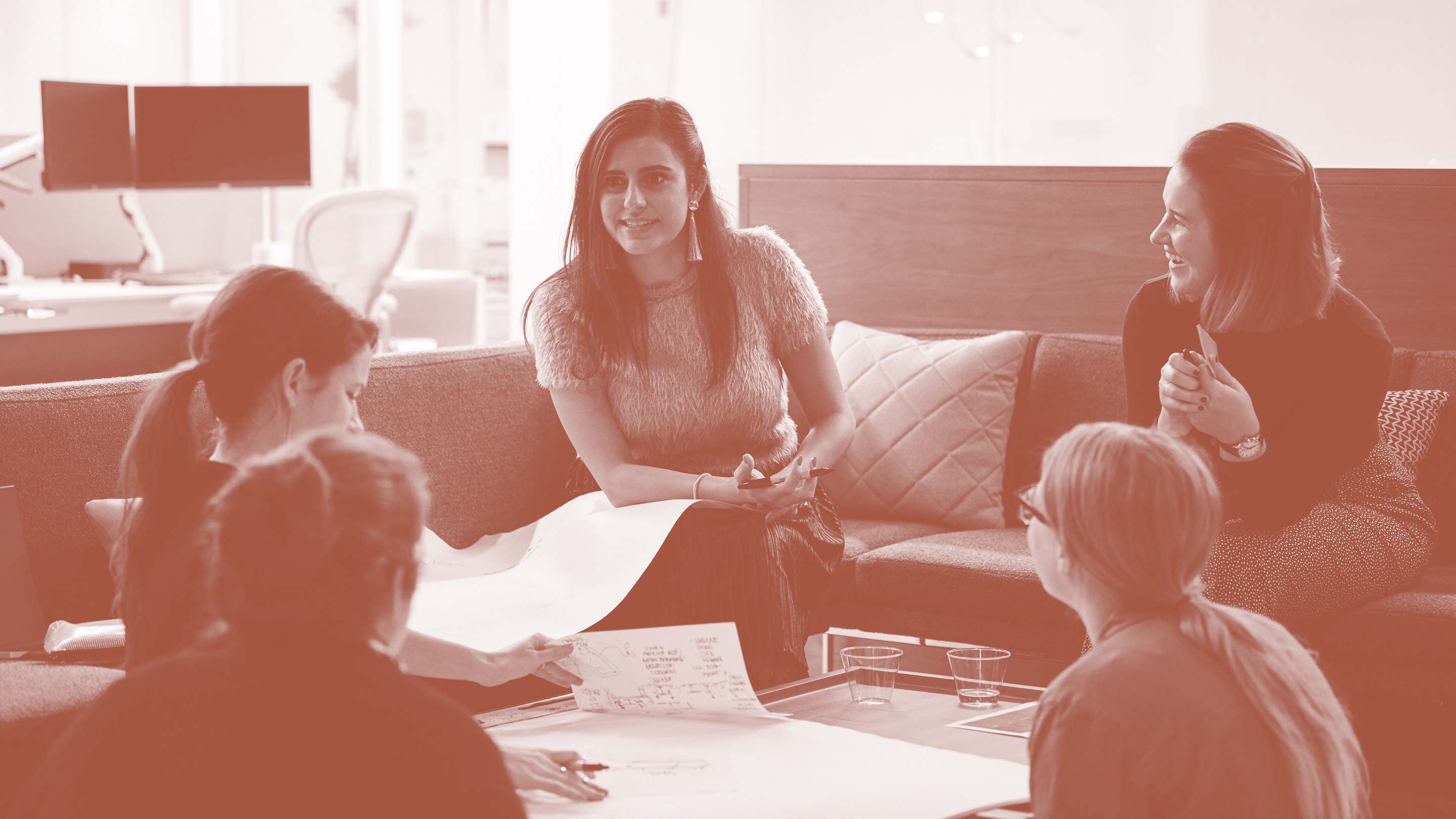 A photo washed in pink. In it, a group of designers chat about the future of work and sketch ideas on a large notepad.