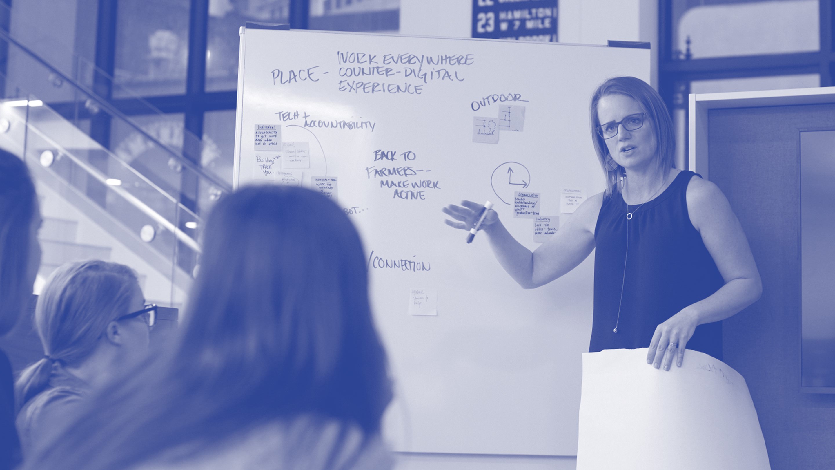 Een foto in blauwe wassing van een persoon die aan een schoolbord staat en een presentatie geeft aan een groepje ontwerpers