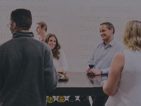 Un grupo de colegas se reúne alrededor de una mesa alta en su oficina para una reunión social después del trabajo. Este tipo de reuniones informales impulsan la innovación.