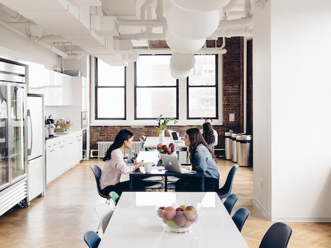 Two women sit and talk inside of an office cafe area full of natural light.