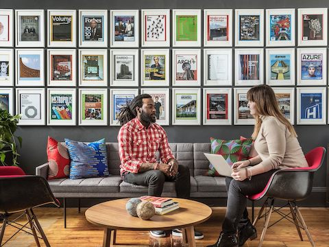 Zwei Mitarbeiter unterhalten sich in einem Lounge-Bereich mit gerahmten Magazin-Covern und Werbeanzeigen an der Wand.