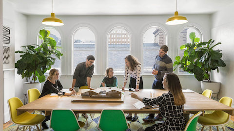 Architecten bij MASS Design werken samen aan een conferentietafel waar groene en gele Eames Shell stoelen aan staan.