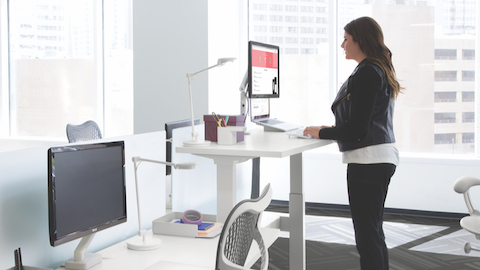 A side view of a woman standing and typing at an adjustable-height desk. Another desk with a chair and adjustable monitor is in the foreground.
